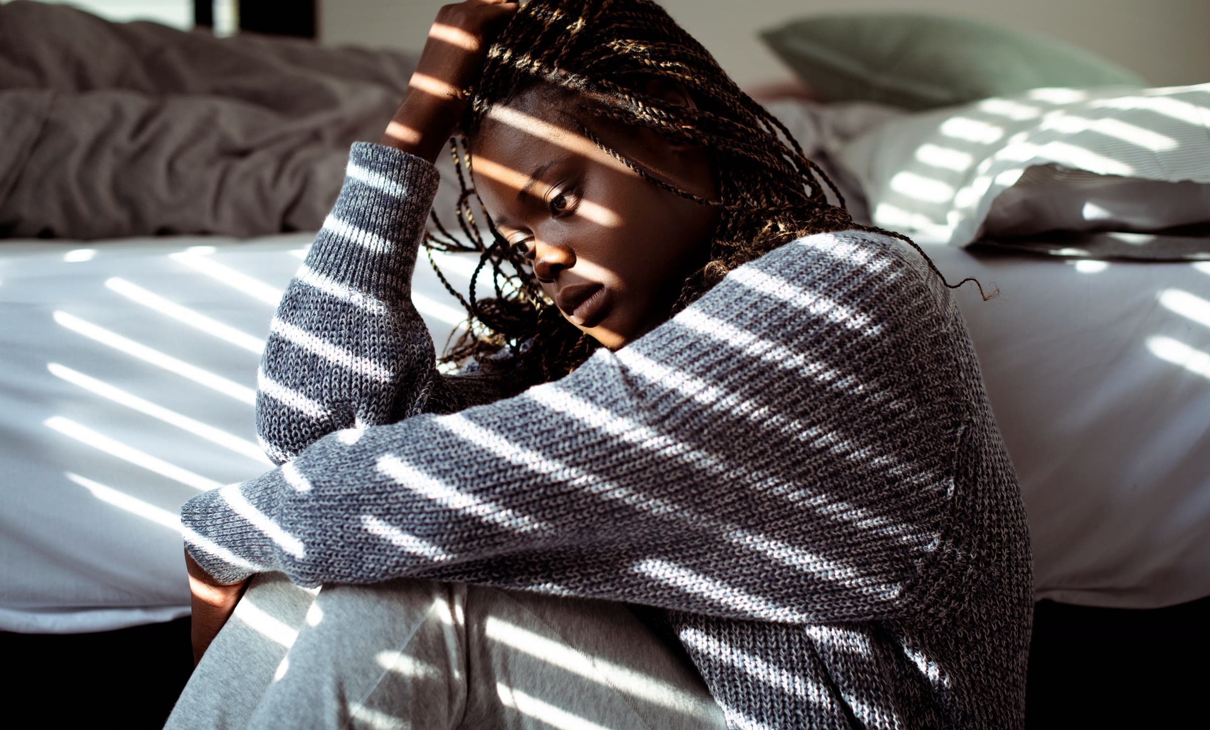 A woman sitting on the floor beside her bed in striped morning light, head resting on her hand, looking exhausted.